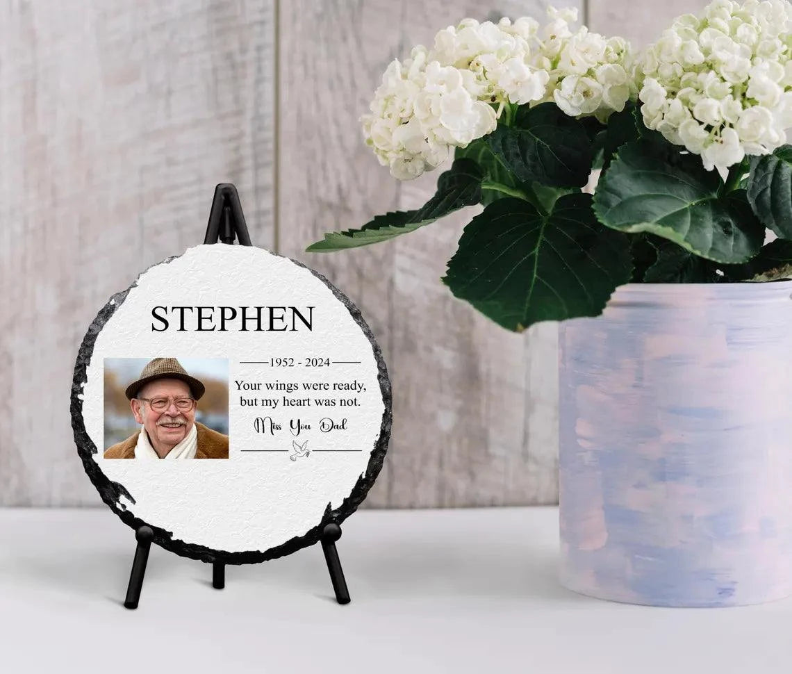 Round personalised memorial slate with full-color photo and glossy finish, displayed on a stand next to flowers.
