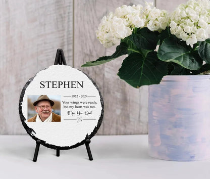 Round personalised memorial slate with full-color photo and glossy finish, displayed on a stand next to flowers.